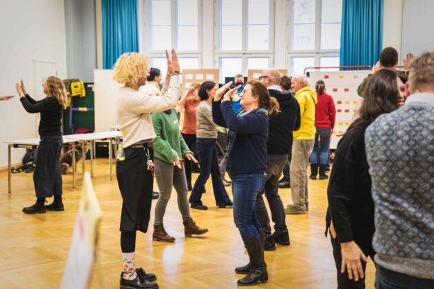 Eine Gruppe von Erwachsenen steht paarweise in einem hellen Raum und nimmt an interaktiven Aktivitäten wie High-Fives und Konversationsübungen teil.