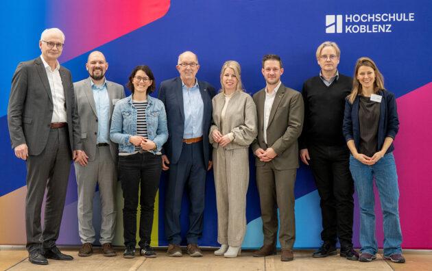 Gruppe von acht Personen steht vor einer farbigen Wand mit dem Schriftzug 'HOCHSCHULE KOBLENZ'