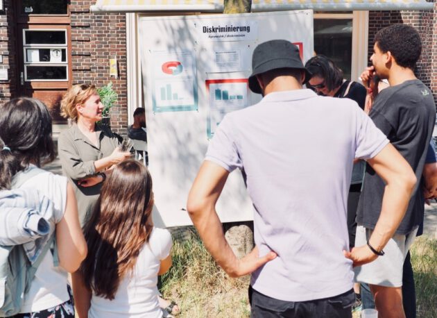 Mehrere Menschen stehen vor einem Whiteboard, auf dem das Wort "Diskriminierung" steht. Die Menschen stehen im Freien und es ist sonnig. Sie scheinen über den Inhalt des Posters zu diskutieren.