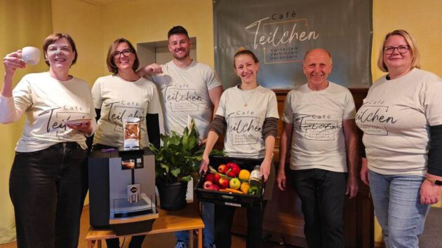 Das Team des Café Teilchen steht lächelnd im Foto. Eine Person hält eine Kiste voller Obst und Gemüse. Im Vordergrund ist eine Kaffeemaschine zu sehen
