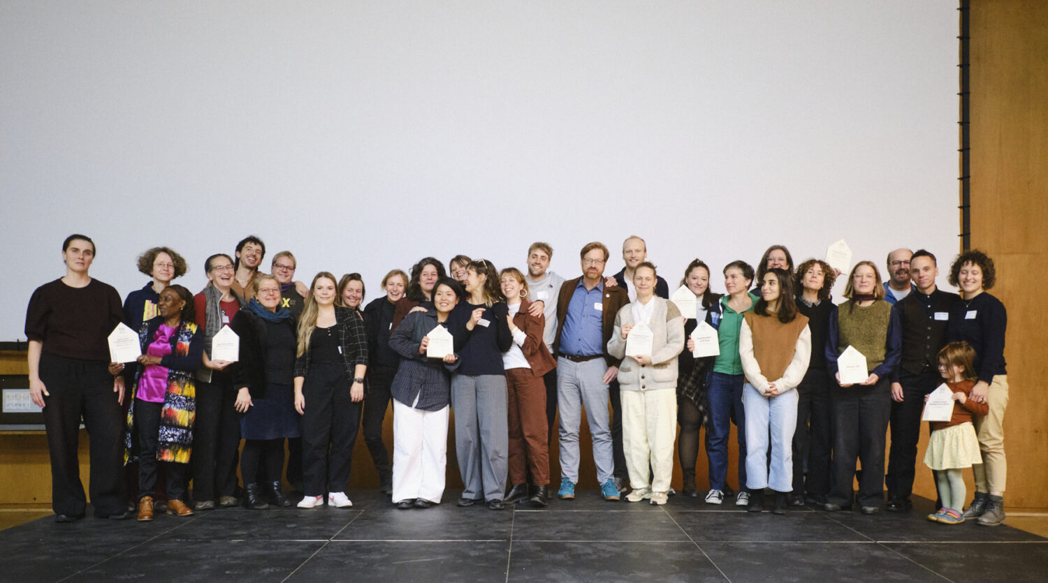 Alle Preisträger*innen des Hans Sauer Preis 2026 sind auf der Bühne in der Aula der Hochschule München zu sehen. Die Personen stehen eng beieinander, lächeln, einige berühren und umarmen sich. Die Stimmung wirkt festlich, ausgelassen und freudig.