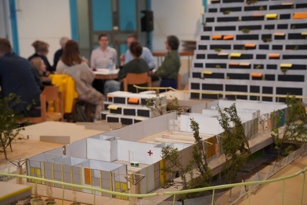 Olykirchen-Werkstatt_Modell-und-Gespraech-1 Das Bild zeigt ein Architekturmodell, das im Vordergrund steht und einen Gebäudekomplex mit mehreren kleinen Gebäuden und Grünflächen darstellt. Im Hintergrund ist eine unscharf dargestellte Gruppe von Personen zu sehen, die an einem Tisch sitzt und sich zu unterhalten scheint. Es handelt sich wahrscheinlich um eine Präsentation oder ein Workshop zu einem Architekturprojekt. Im Hintergrund ist ein weiteres, größeres Architekturmodell zu erkennen - ein Hochhaus mit vielen kleinen, farbig markierten Balkonen.