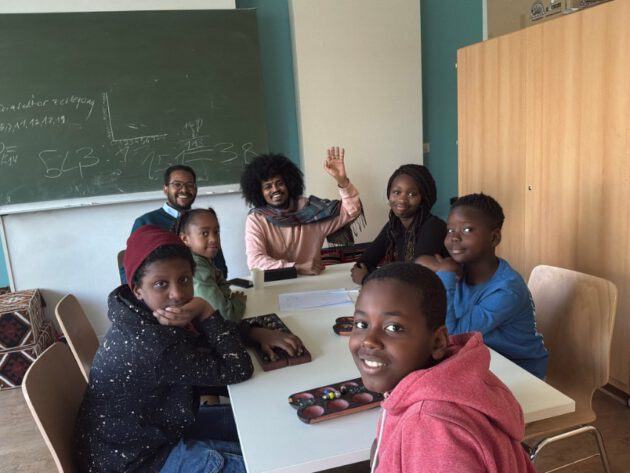 Sechs Kinder und zwei Erwachsene sitzen um einen rechteckigen Tisch in einem Klassenzimmer. Die Kinder tragen Freizeitkleidung in verschiedenen Farben und blicken freundlich in Richtung Kamera. Eine der erwachsenen Personen hebt die Hand zum Gruß. Im Hintergrund steht eine grüne Tafel mit handschriftlichen Notizen und Diagrammen. Rechts befindet sich ein heller Schrank, links sind bunte Sitzkissen zu sehen. Die Atmosphäre wirkt offen und einladend.
