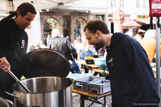 Zwei Männer in Kapuzenpullovern stehen an einem großen dampfenden Kochtopf auf einem Markt mit Tischen und Menschen im Hintergrund