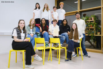 Die jungen Macher*innen des Umwelt-Magazins sitzen und stehen auf gelben Stühlen in einem Workshop-Raum und blicken in Richtung Kamera. Eine sitzende Person in der Mitte der Gruppe hält die Urkunde des Hans Sauer Preis in den Händen.