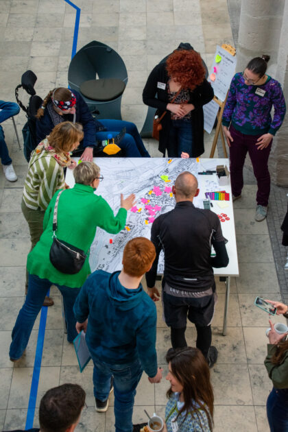 Mehrere Menschen stehen um einen Tisch herum, auf dem eine Karte ausgebreitet ist. Die Karte ist mit Post-Its beklebt. Die Menschen sind von oben aufgenommen. Sie deuten auf unterschiedliche Punkte der Karte und befinden sich im Gespräch dazu.
