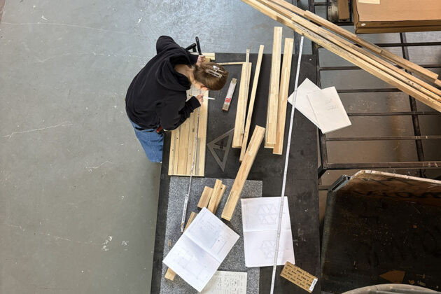 Von oben sichtbare Person mit Zopf, die an einem Tisch mit Holzleisten, Zeichnungen und Messwerkzeugen arbeitet